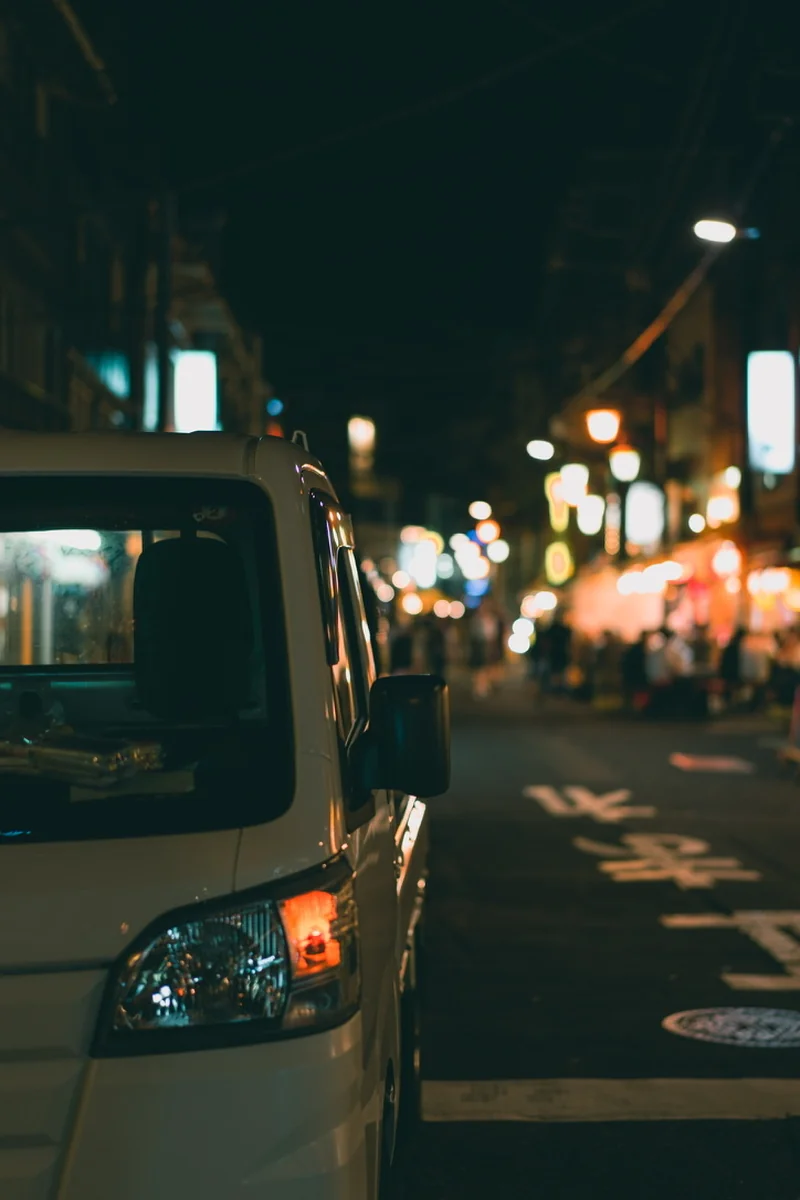 A small white truck parked beside a street lit by shop signs.